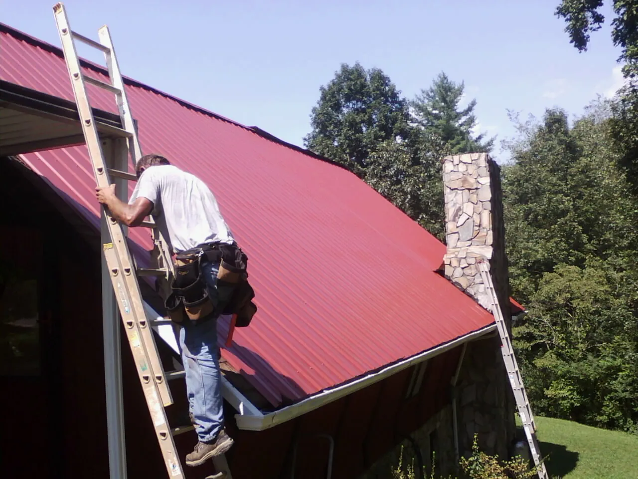 Roofer on a ladder beside a red metal roof and stone chimney — Soffit & Fascia Repair by Roofing Brothers in East Sparta